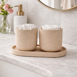 Puede incluir: Un juego de dos recipientes cilíndricos de color beige moteado sobre una bandeja ovalada. Un recipiente está lleno de bastoncillos de algodón blancos y el otro contiene discos de algodón blancos. Los recipientes y la bandeja tienen un acabado mate.