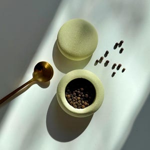 May include: A light green ceramic pepper grinder with a matching lid. The grinder is filled with black peppercorns and is sitting on a white surface. A gold spoon is also in the image.