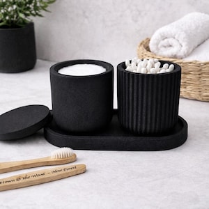 May include: A set of two matte black cylindrical containers on an oval tray. One container has a smooth surface and holds cotton rounds, while the other has a ribbed texture and holds cotton swabs. A matching black lid sits beside the tray.
