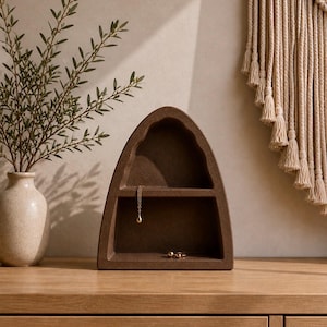 May include: A brown, arched jewelry organizer with two compartments. A gold necklace and small rings are displayed inside. The organizer sits on a wooden surface, with a vase of greenery to the left and a macrame wall hanging in the background.
