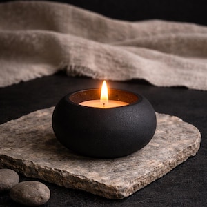 Pode incluir: Um suporte de vela redondo, preto fosco, com uma superfície texturizada contendo uma vela de chá acesa. A chama emite um brilho quente e brilhante do centro do recipiente circular escuro, que repousa sobre uma superfície de pedra plana.