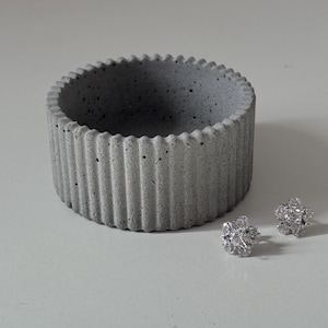 May include: A grey concrete trinket dish with a ribbed design. The dish is round and has a slightly raised lip. Two small silver earrings are sitting on the surface next to the dish.