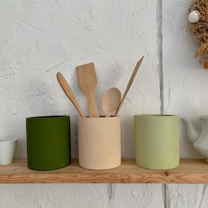 Pu&ograve; includere: Tre portautensili in ceramica di colore verde, beige e verde chiaro con una finitura macchiettata. Cucchiai e spatole di legno sono posizionati nei portautensili.