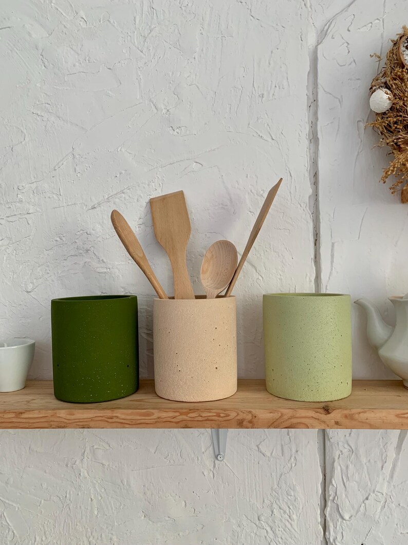 Pu&ograve; includere: Tre portautensili in ceramica di colore verde, beige e verde chiaro con una finitura macchiettata. Cucchiai e spatole di legno sono posizionati nei portautensili.