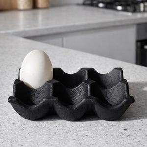 May include: A black, six-egg holder with a single white egg in the first slot. The holder has a wavy design and is made of a matte material. The kitchen setting includes a countertop and a blurred background.