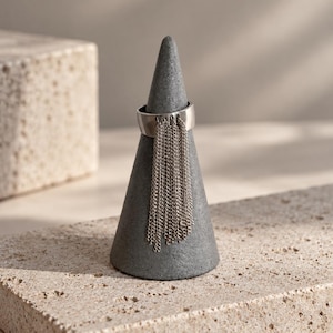May include: A silver-toned metal ring featuring a wide band with a series of delicate, dangling chain fringe elements attached to the front. The ring is displayed on a dark gray, cone-shaped stand, showcasing the movement of the metallic chains.