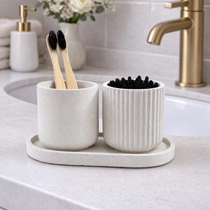 Puede incluir: Un juego de baño sobre una encimera con dos recipientes cilíndricos y una bandeja. Un recipiente contiene dos cepillos de dientes de bambú con cerdas negras, y el otro bastoncillos de algodón negros. El conjunto es de color blanco roto moteado.
