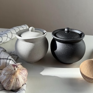May include: Two ceramic lidded jars, one light gray and one black, sit side-by-side. The jars have a round shape with a small knob on the lid. A bulb of garlic and a wooden spoon are visible near the jars. A checkered cloth is partially visible.