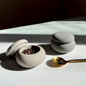 May include: Two grey stone containers with lids, one filled with black peppercorns, and a gold spoon with peppercorns on a white surface.