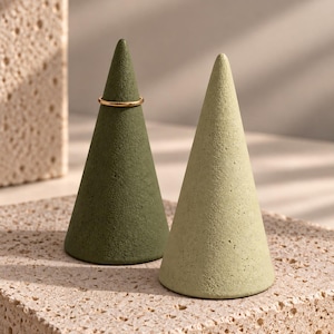 May include: Two cone-shaped ring holders with a textured matte finish. One cone is dark olive green and the other is a lighter sage green. A thin, plain gold-colored metal ring is placed around the top section of the dark olive green cone.