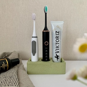 May include: A green concrete toothbrush holder with three slots. Two electric toothbrushes, one white and one black, are in the holder. A tube of toothpaste with the brand name "VIKTORIZ" is in the third slot.