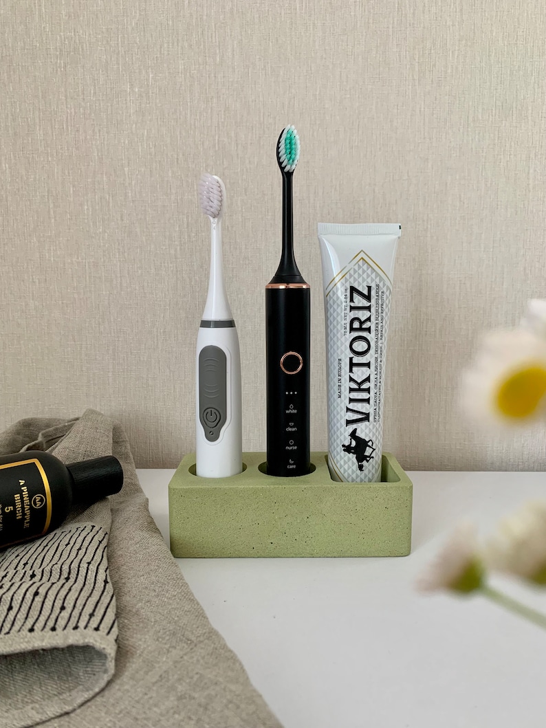 May include: A green concrete toothbrush holder with three slots. Two electric toothbrushes, one white and one black, are in the holder. A tube of toothpaste with the brand name "VIKTORIZ" is in the third slot.