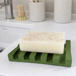 Pode incluir: Uma barra de sabão de cor clara repousa sobre uma saboneteira verde com um design ranhurado. A saboneteira está sobre uma bancada branca, com um porta-escovas de dentes, escovas de dentes e um dispensador de sabão no fundo.