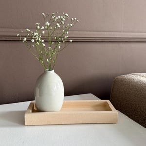 Puede incluir: Un jarrón de cerámica blanco de forma redondeada contiene delicadas flores blancas. El jarrón está sobre una bandeja rectangular de color beige claro. Los objetos están sobre una superficie blanca, con una pared marrón al fondo.