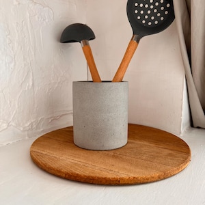 Pu&ograve; includere: Un portautensili in cemento grigio con due utensili da cucina in silicone nero, un mestolo e una schiumarola, con manici in legno.