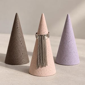 May include: A silver ring with a dangling chain fringe feature displayed on a pale pink concrete cone. The ring is positioned between a dark brown concrete cone on the left and a light lavender concrete cone on the right, all with textured surfaces.