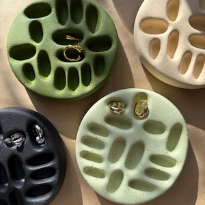 Könnte beinhalten: Vier kreisförmige Schmucktabletts in Grün-, Creme- und Schwarztönen. Jedes Tablett hat eine Reihe von ovalen Aussparungen. Goldene und silberne Ohrringe werden in den Tabletts ausgestellt. Die Tabletts sind auf einer braunen Oberfläche angeordnet.