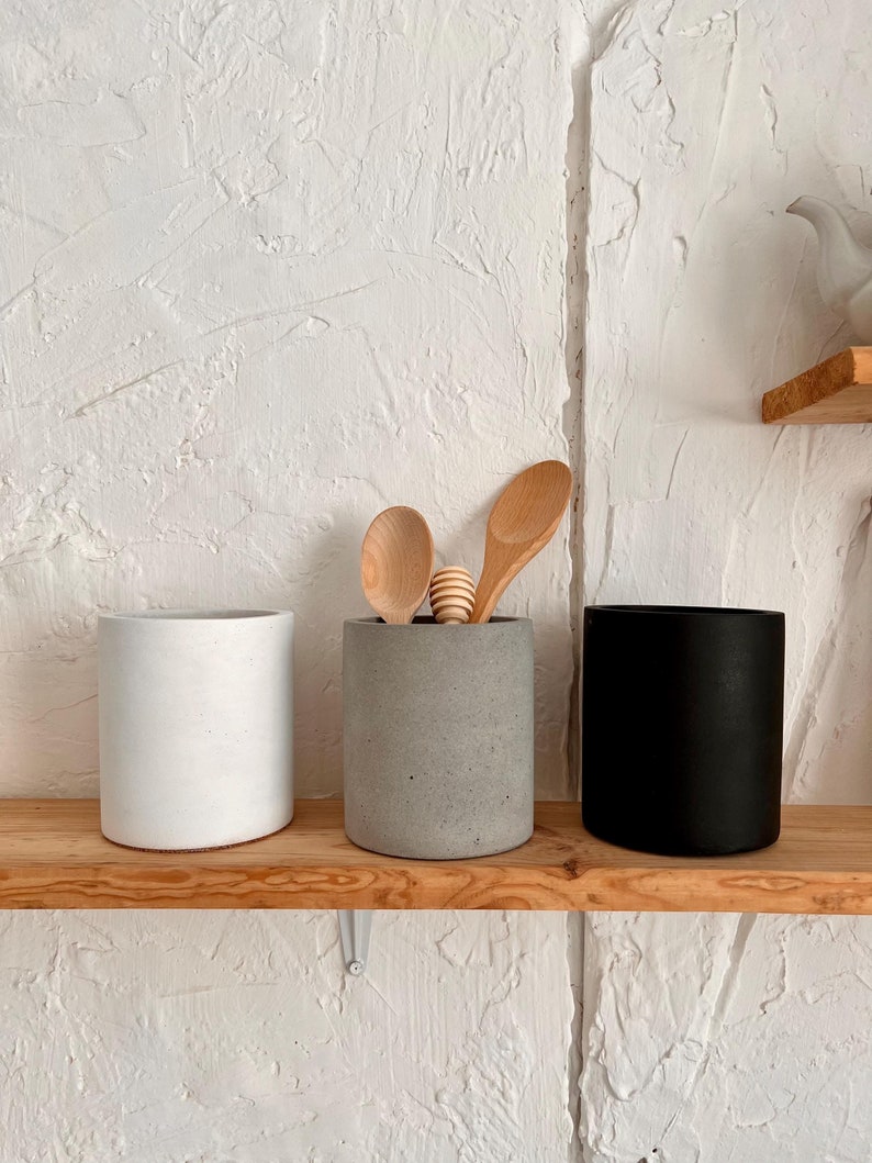 Pu&ograve; includere: Tre portautensili cilindrici in cemento di colore bianco, grigio e nero su uno scaffale in legno. Due cucchiai di legno sono nel portautensili grigio.
