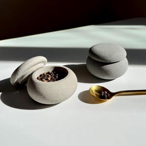 Op de afbeelding: Twee grijze stenen containers met deksels, een gevuld met zwarte peperkorrels, en een gouden lepel met peperkorrels op een wit oppervlak.