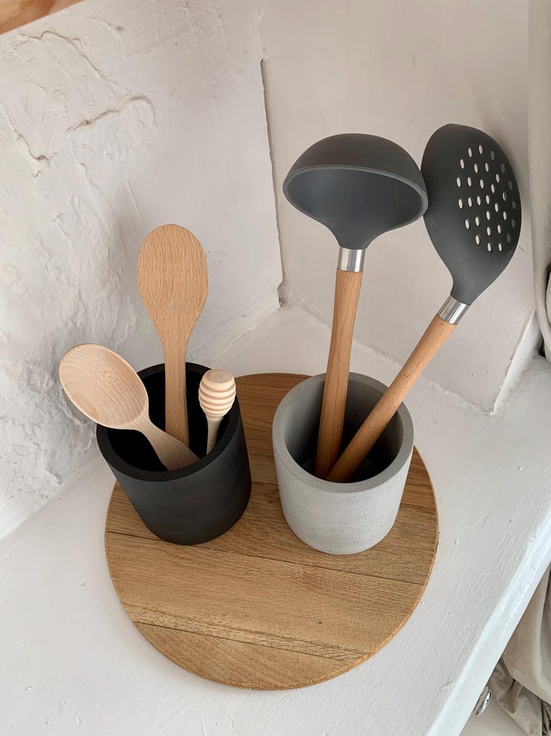 Pu&ograve; includere: Un vassoio rotondo in legno con due portautensili in cemento. Un portautensili &egrave; nero e l'altro &egrave; grigio. I portautensili contengono cucchiai di legno e una mestolina e una paletta forata in silicone grigio.