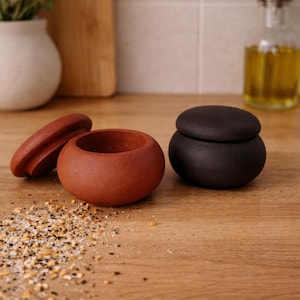 Op de afbeelding: Een terracotta-kleurige vijzel en stamper met een deksel, en een zwarte keramische container met een deksel, staan op een houten oppervlak. Kruiden zijn verspreid over het oppervlak. Een snijplank, olijfolie en een vaas met groen staan op de achtergrond.