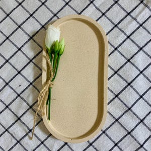 May include: A beige oval tray with a speckled texture. A single white flower with green stems and leaves is placed on the tray.