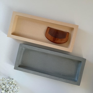 Pode incluir: Duas bandejas retangulares de concreto, uma na cor bege claro e a outra na cor cinza. A bandeja bege tem um pente de madeira dentro.