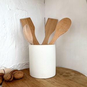 Pu&ograve; includere: Un portautensili in ceramica bianca con tre cucchiai e spatole di legno al suo interno. Il portautensili &egrave; appoggiato su un tagliere di legno.