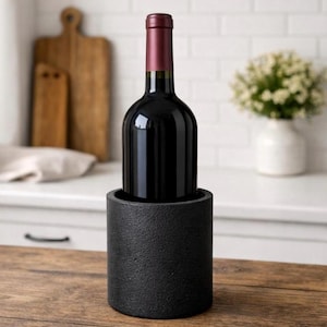 Puede incluir: Una botella de vino tinto con cuello burdeos se coloca en un enfriador de vino negro cilíndrico. El enfriador tiene una superficie texturizada y descansa sobre una mesa de madera. El fondo incluye una encimera de cocina blanca y una tabla de cortar.