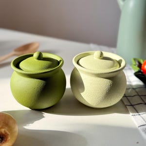 Op de afbeelding: Twee kleine, ronde keramische potten met deksel in groen- en geelgroentinten. De potten hebben een getextureerd oppervlak en een kleine knop bovenop het deksel. Op de achtergrond staat een lichtgroene kan.