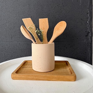 Puede incluir: Un porta utensilios de hormigón beige con cuatro utensilios de cocina de madera, incluyendo una cuchara, una espátula y dos cucharas de madera, y una ramita de flores blancas.