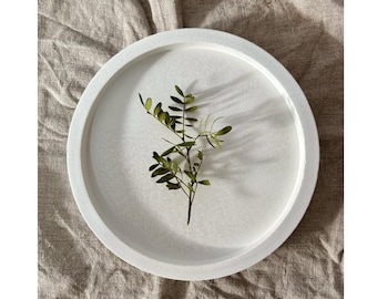 May include: A round, white concrete tray with a raised edge, holding a sprig of green leaves. The tray sits on a beige linen surface, creating a minimalist and natural aesthetic. The design is simple and elegant.