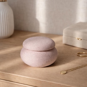 May include: A light pink, stone-like jewelry box with a lid, sitting on a wooden surface. A gold necklace and ring are also visible. The box has a round shape and a textured surface, suggesting a natural aesthetic.