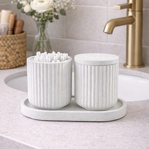 May include: A set of two cylindrical, ribbed, speckled white containers made of a stone-like material, one with a lid and one filled with cotton swabs, both resting on an oval tray of the same matching material and speckled white finish.