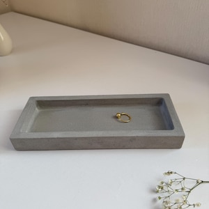 May include: A rectangular, gray concrete tray with a gold ring. The tray is a simple, minimalist design, perfect for holding jewelry or small items. The gold ring adds a touch of elegance to the otherwise industrial aesthetic.