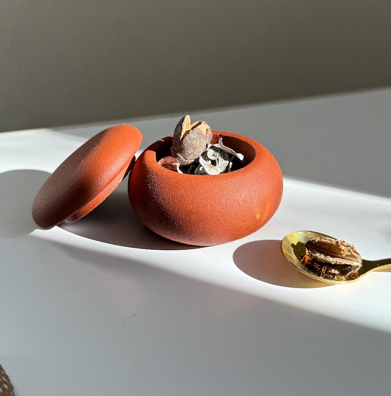 Op de afbeelding: Een terracotta keramische container met een deksel, gevuld met gedroogde botanicals. De container staat op een wit oppervlak naast een gouden lepel met gedroogde botanicals.