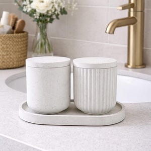 Può includere: Due barattoli in ceramica bianca con coperchi su un vassoio ovale. Uno ha una finitura maculata, l'altro dettagli a coste verticali. Un rubinetto dorato e un vaso di fiori bianchi sono sullo sfondo. Arredamento bagno.