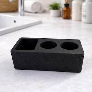 Puede incluir: Un organizador de baño rectangular negro con tres compartimentos. El organizador tiene dos agujeros circulares y un compartimento cuadrado. Está hecho de un material texturizado y se encuentra sobre una superficie blanca.