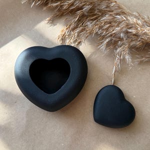 Può includere: Due oggetti neri a forma di cuore. Uno è un cuore pieno e l'altro è un contenitore a forma di cuore. Gli oggetti sono su una superficie dai toni neutri con erba ornamentale secca sullo sfondo. Gli oggetti sono probabilmente decorativi.