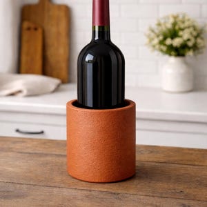 Puede incluir: Un enfriador de botellas de vino de color terracota sostiene una botella de vino oscuro con una tapa roja. El enfriador cilíndrico tiene una superficie texturizada y está sobre una mesa de madera. Una tabla de cortar y flores están en el fondo.