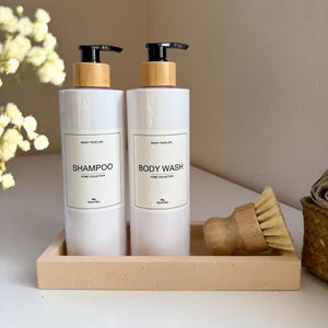 Op de afbeelding: Twee witte shampoo- en douchegeldispensers met zwarte pompen en bamboekleurige doppen staan op een lichtbruin dienblad. De dispensers hebben labels met de tekst "Enjoy Your Life" en "Home Collection."