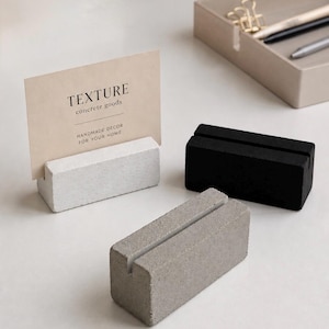 May include: Three rectangular concrete card holders in white, gray, and black. The white holder supports a beige card with the text "TEXTURE concrete goods HANDMADE DECOR FOR YOUR HOME" printed in black. Each holder features a narrow slot on top.