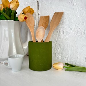 Pu&ograve; includere: Un portautensili in ceramica verde macchiata con quattro cucchiai e spatole di legno all'interno. Il portautensili &egrave; su una superficie bianca con un'anfora bianca e tulipani gialli sullo sfondo.