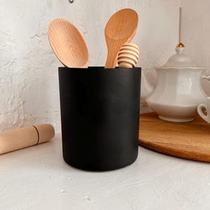 Pu&ograve; includere: Un portautensili nero con cucchiai di legno e un mestolo per il miele all'interno. Il portautensili &egrave; su un bancone bianco.