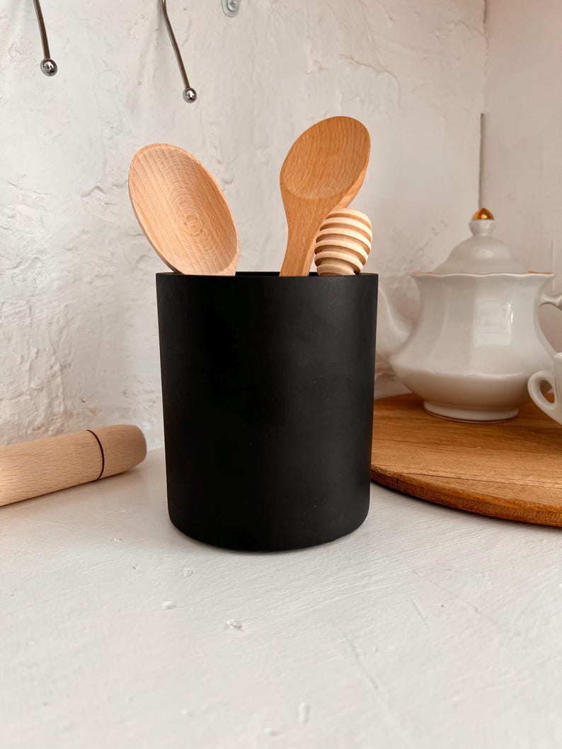 Pu&ograve; includere: Un portautensili nero con cucchiai di legno e un mestolo per il miele all'interno. Il portautensili &egrave; su un bancone bianco.