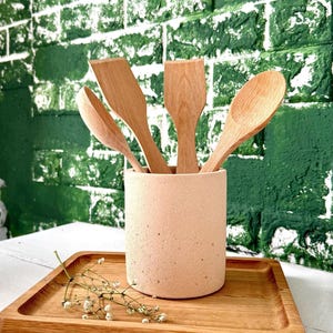 Puede incluir: Un juego de utensilios de cocina de madera, que incluye una cuchara y espátulas, en un soporte cilíndrico beige. El soporte está sobre una bandeja de madera con una ramita de flores blancas. El fondo es una pared de ladrillos verdes.