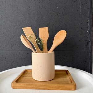 Pu&ograve; includere: Un portautensili in cemento beige con quattro utensili da cucina in legno al suo interno. Il portautensili &egrave; posto su un vassoio di legno su un tavolo bianco.