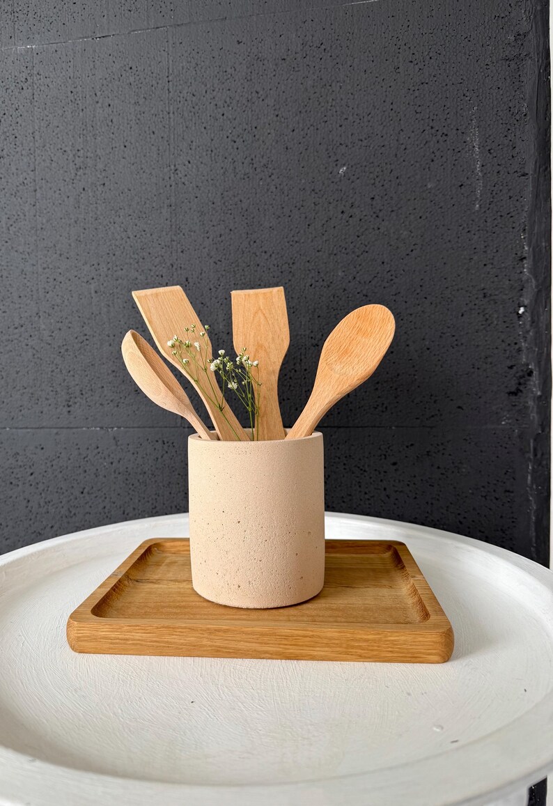 Pu&ograve; includere: Un portautensili in cemento beige con quattro utensili da cucina in legno al suo interno. Il portautensili &egrave; posto su un vassoio di legno su un tavolo bianco.