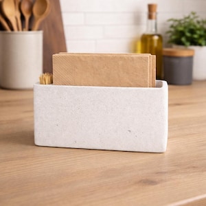 Può includere: Un portatovaglioli rettangolare in cemento grigio chiaro riempito con tovaglioli di carta marrone e un piccolo mazzetto di stuzzicadenti in legno. Il supporto è appoggiato su un piano di lavoro in legno, con utensili da cucina e olio d'oliva sullo sfondo.