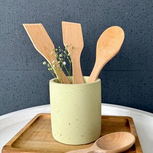 Pu&ograve; includere: Un set di utensili da cucina in legno, tra cui un cucchiaio, una spatola e un cucchiaio pi&ugrave; piccolo, sono esposti in un portautensili in cemento verde chiaro. Il portautensili &egrave; appoggiato su un vassoio di legno.